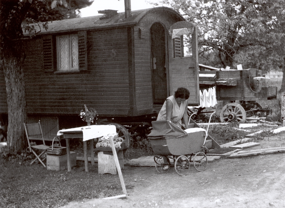Black-and-white photograph of a woman with a child in front of a caravan. The child is in a pram. In the background, laundry is drying, and in the foreground there is a small table with a bouquet of flowers.