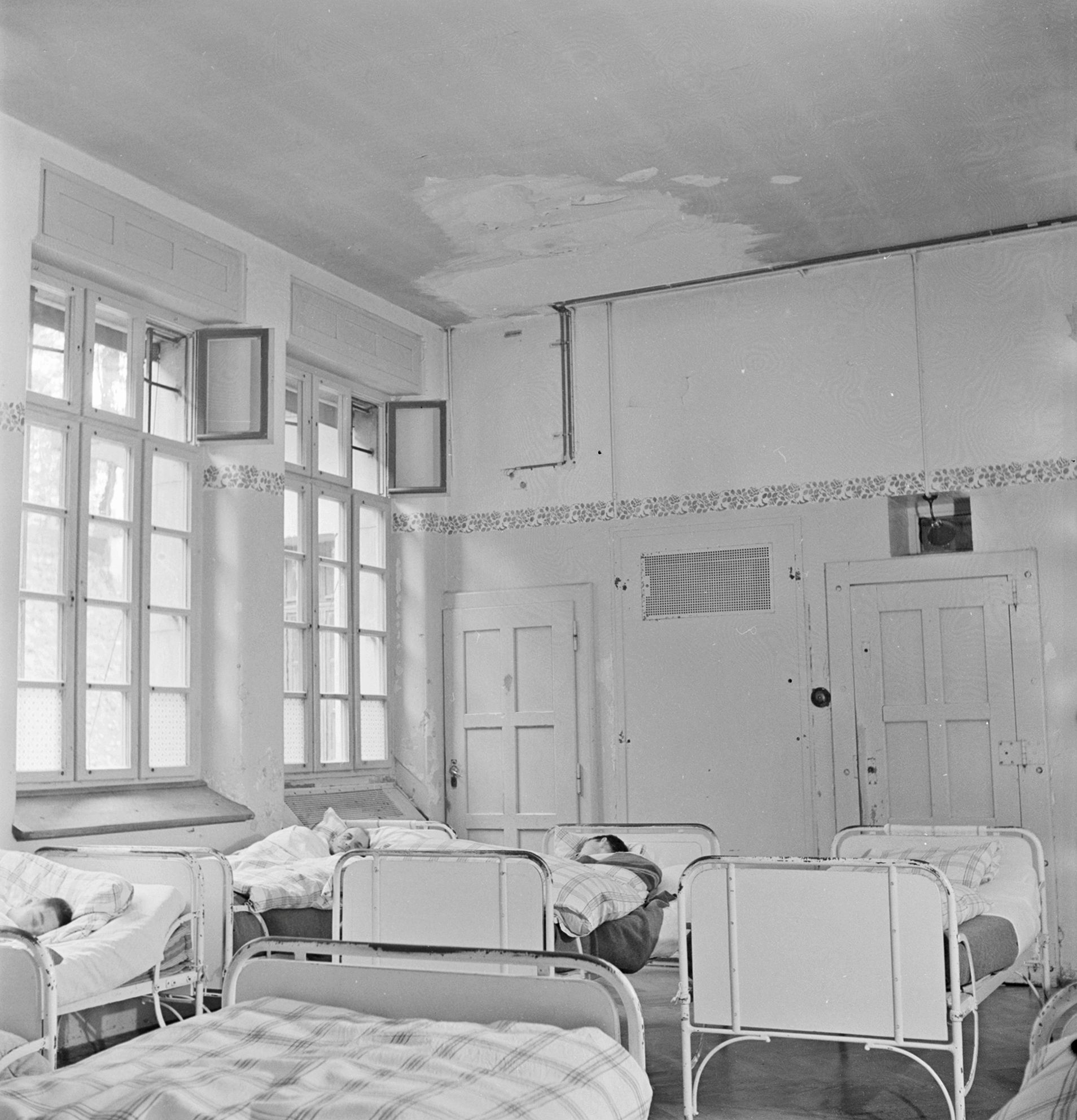Black-and-white photograph of a room in a psychiatric clinic around 1948. Several metal beds can be seen, three of which have people lying in them with their heads turned away.