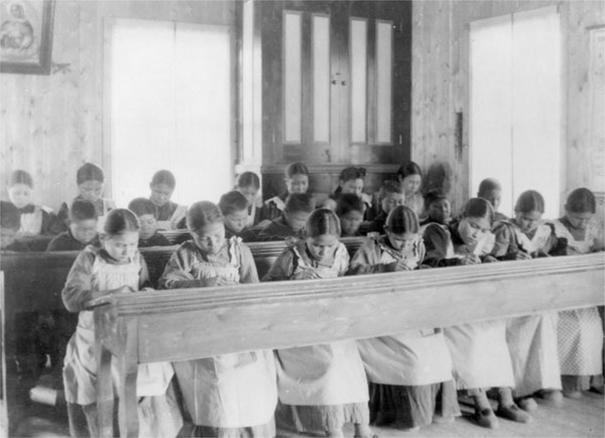 Black-and-white photograph of a classroom with wooden school desks, in which young girls are sitting and writing. This is a so-called ‘residential school’ (reform schools) for indigenous children in Canada.