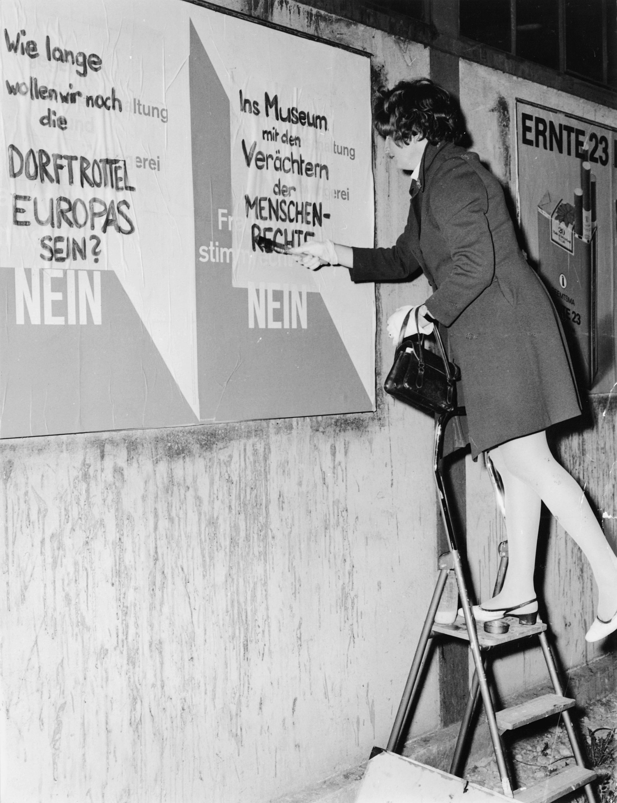 Black-and-white photograph of a woman standing on a ladder and covering up a poster by opponents of women's suffrage. The text on her posters reads: ‘How long do we want to remain the village idiots of Europe?’ and ‘Put the despisers of human rights in a museum’. The photograph was probably taken during the ‘March on Bern’ in 1969.
