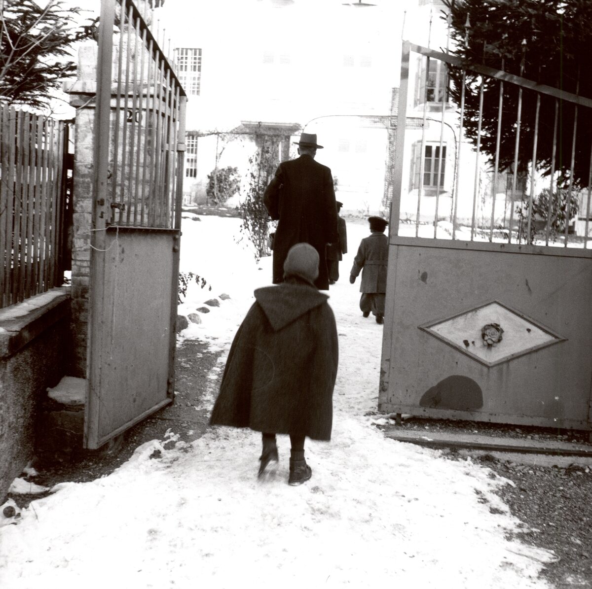 The black-and-white photograph shows a winter scene in front of a building, presumably an institution for Yenish children. In the foreground, a child wearing a coat and a hat is seen walking towards an open gate. Behind the child, Dr Alfred Siegfried, wearing a hat and coat, and two other children are walking towards the building. The ground is covered with snow, and the gate opens onto a courtyard with a multi-storey house in the background.