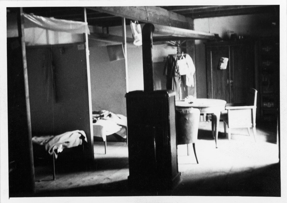 Black-and-white photograph of a room with a heater, beds and a table. Presumably accommodation for seasonal workers, 1970. The image is from the archives of the newspaper Vorwärts.