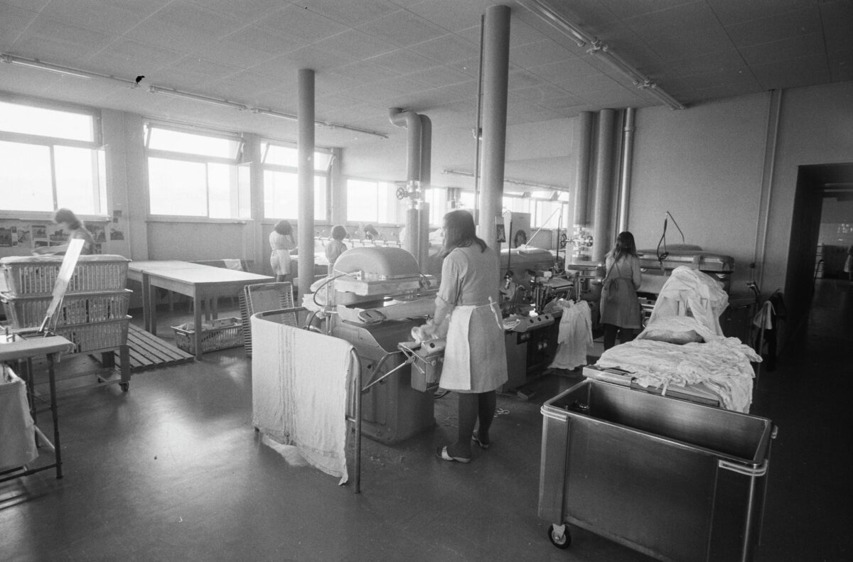 Black-and-white photograph of the laundry room at Hindelbank women's prison. Four women are seen from behind as they work on the ironing machines. 1967.