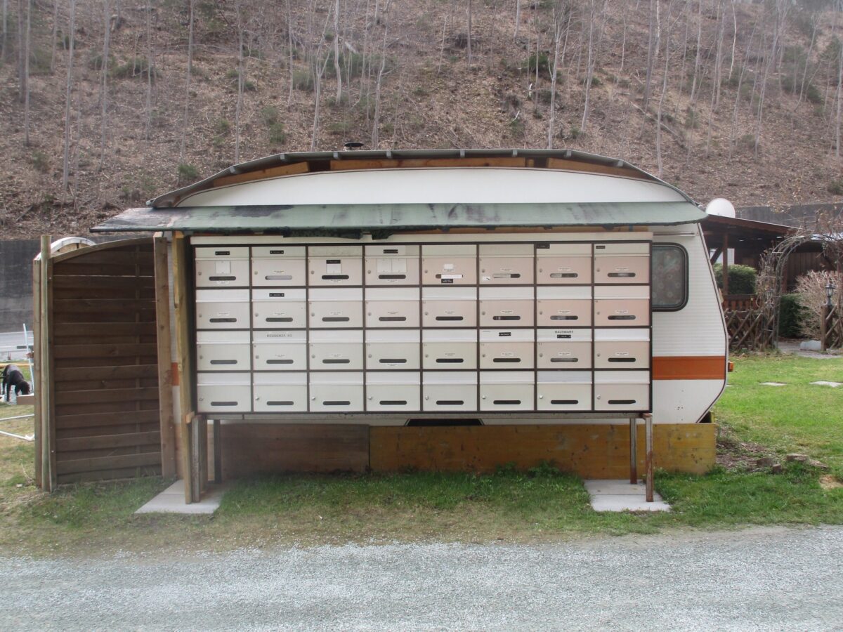 Colour photograph of 32 letterboxes in front of a caravan. These are the letterboxes of the tenants of the Rania campsite in the municipality of Zillis-Reischen. The photograph was taken in April 2016 by Willi Wottreng. The Rania campsite is currently the only site in Switzerland managed by the Yenish people.