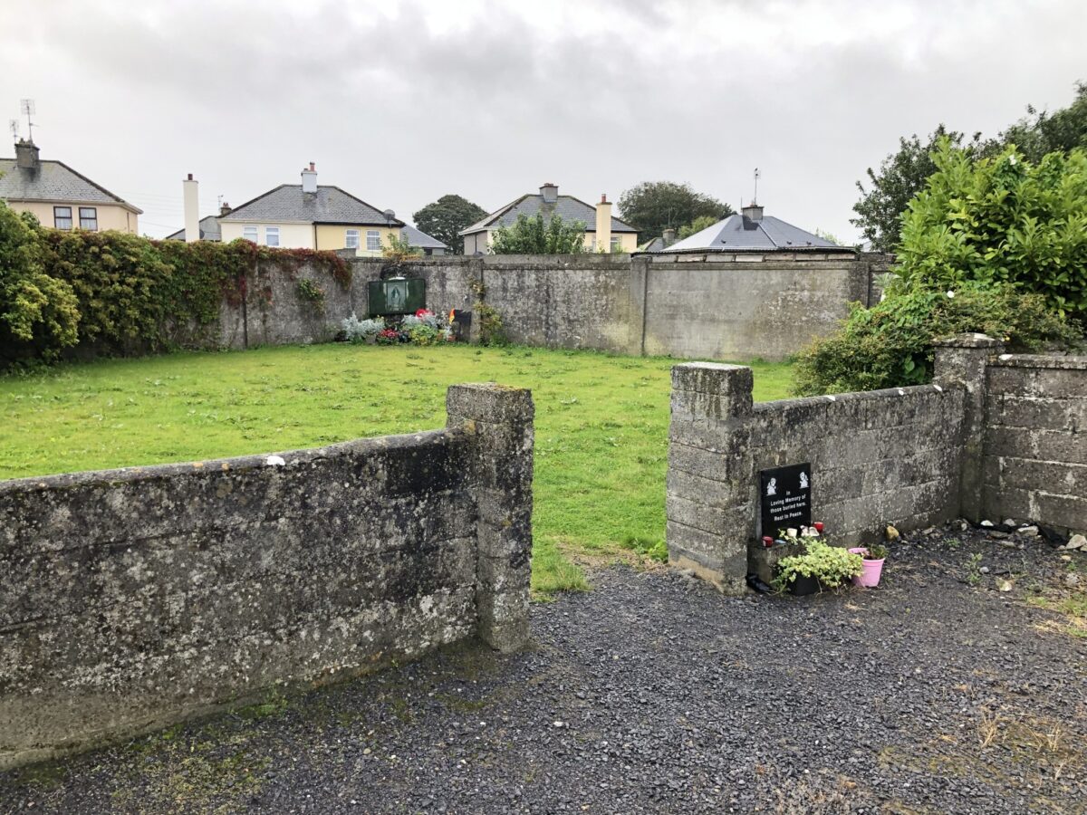 Colour photograph of a fenced courtyard with stone walls. View from the entrance. Flowers and memorial plaques can be seen at the entrance and on the opposite side. This is a memorial to the children who died at the Bon Secours Mother and Baby Home in Tuam, Galway.