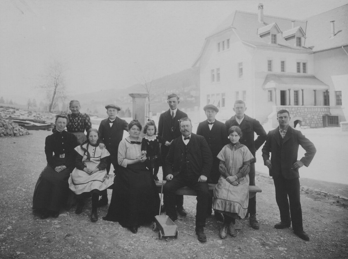 Black-and-white photograph of women, men and children posing for a photo, standing and sitting on a bench in elegant clothing. They are probably staff and ‘pupils’ of the reformatory for boys in Sonvilier. The photograph was shown at the National Exhibition in Bern in 1914 and documented the work of the Directorate for Welfare of the Canton of Bern.