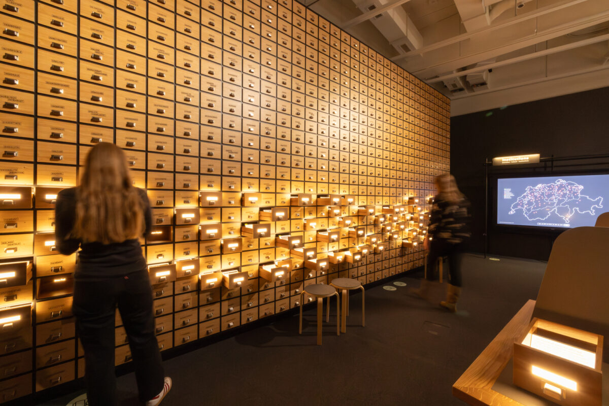 Eine Wand voller Schubladen, einzelne sind beleuchtet. Zwei Personen schauen sich diese an.