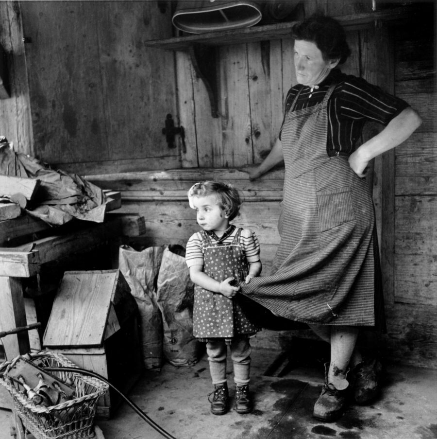 Black-and-white photograph of a small child clinging to the hem of an adult woman's skirt. The child is a foster child and the woman is his foster mother. The photograph was taken by Walter Studer in Emmental in 1954. The walls of a wooden house can be seen in the background, and a doll's pram stands in the foreground.