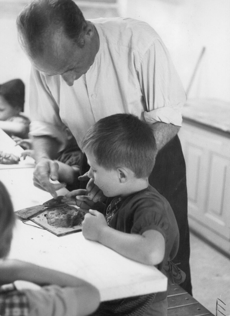 Black-and-white photograph of an adult man and children at a table. The man is standing, while the children are sitting at the table doing crafts. They are the foster children and foster father of a large foster family around 1950.