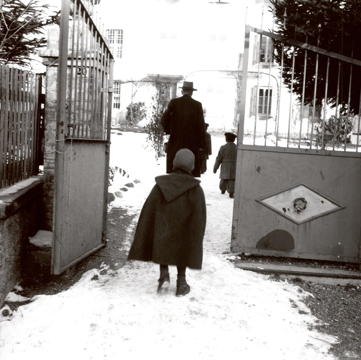The black-and-white photograph shows a winter scene in front of a building, presumably an institution for Yenish children. In the foreground, a child wearing a coat and a hat is seen walking towards an open gate. Behind the child, Dr Alfred Siegfried, wearing a hat and coat, and two other children are walking towards the building. The ground is covered with snow, and the gate opens onto a courtyard with a multi-storey house in the background.