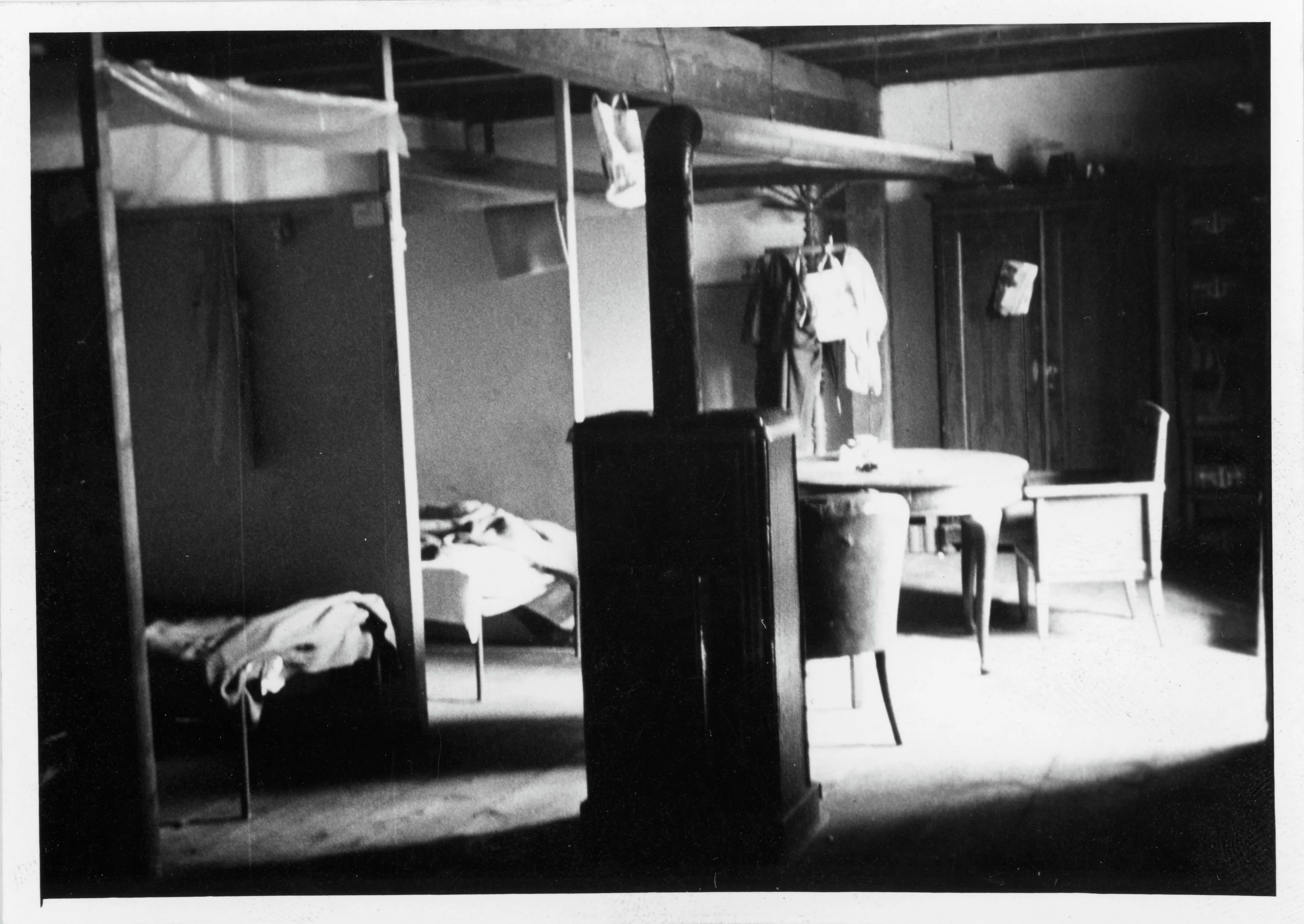 Black-and-white photograph of a room with a heater, beds and a table. Presumably accommodation for seasonal workers, 1970. The image is from the archives of the newspaper Vorwärts.