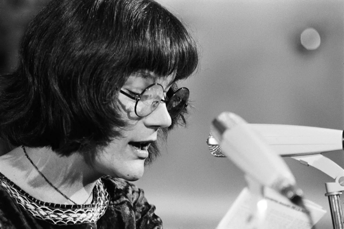 Black-and-white photograph of a woman standing in front of several microphones at a lectern. The woman is Mariella Mehr, Swiss writer (1949–2022).