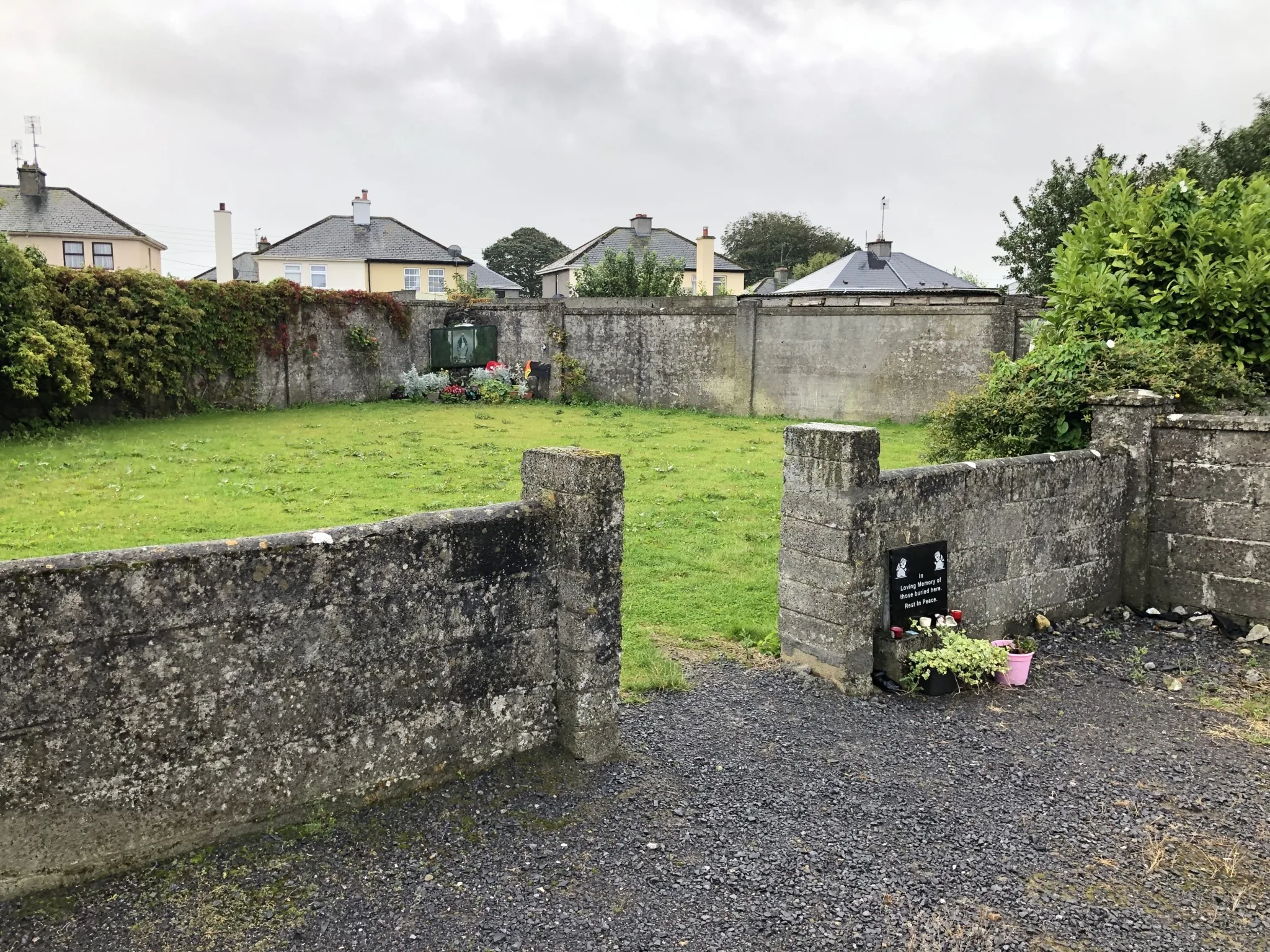 Colour photograph of a fenced courtyard with stone walls. View from the entrance. Flowers and memorial plaques can be seen at the entrance and on the opposite side. This is a memorial to the children who died at the Bon Secours Mother and Baby Home in Tuam, Galway.